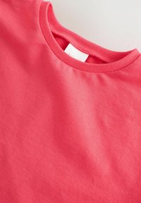 Camiseta de manga corta en coral brillante, confeccionada con una suave mezcla de algodón. Cuenta con cuello redondo y textura lisa, con una etiqueta en el cuello.