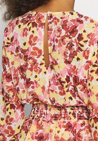 Abito floreale con un motivo in rosa, giallo e rosso. Presenta una schiena a occhiello, maniche lunghe e una vita arricciata per una vestibilità su misura.