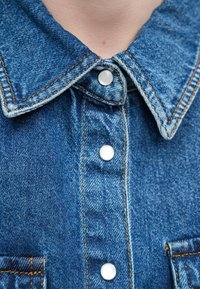 Πουκάμισο από τζιν με μυτερό γιακά, ανοιχτό μπλε ύφασμα, ασημένια κουμπώματα τύπου snap, πορτοκαλί ραφές και μπροστινή τσέπη με υφή.