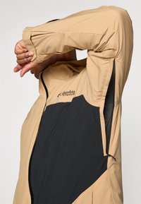 Beige og svart Columbia Titanium-jakke med glidelås foran, sideventiler og elastiske mansjetter, laget av glatt og lett stoff.