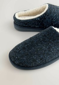 Tmavě modré pantofle vyrobené z texturované tkaniny se měkkým bílým podšívkou, zakulacenou špičkou a pevným gumovým podešví.