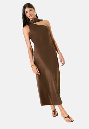 Frau trägt ärmelloses braunes asymmetrisches Maxikleid mit Neckholder, Sandalen sowie goldene Ohrringe und Armband, posiert vor weißem Hintergrund.