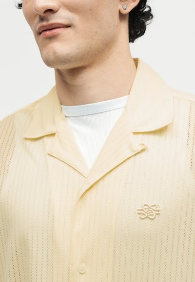 Κοντινό πλάνο ενός ατόμου που φοράει μια κρεμ πουκάμισο με κουμπιά, υφή και κεντημένο λογότυπο, πάνω από λευκό ριμπ μπλουζάκι με στρογγυλή λαιμόκοψη.