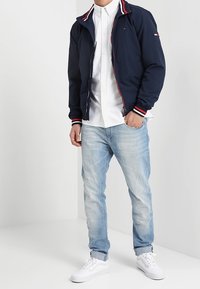 Navy zip-up jack met een rood en wit gestreept kraag, witte button-up blouse, lichtblauwe spijkerbroek en witte sneakers. Casual outfit.