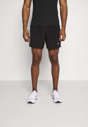 Mann trägt schwarze Sportshorts, schwarzes Shirt und weiße Laufschuhe, steht vor einem schlichten hellen Hintergrund.