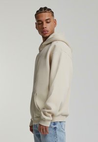 Felpa con zip beige in materiale morbido, caratterizzata da un ampio cappuccio, tasca frontale con busta e polsini a costine. Indossata con jeans azzurri chiari.