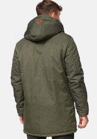 Mann in einer olivgrünen Winterjacke mit Kapuze, von hinten vor einem weißen Hintergrund betrachtet.