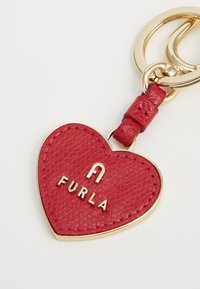 Crveni kožni privjesak u obliku srca s teksturiranom površinom, zlatnim hardverom i urezanim logotipom "FURLA" na sredini.