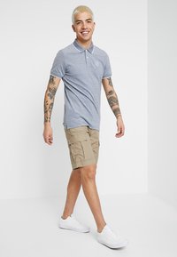 Grå polo-shirt med krave og logo, parret med khaki cargo shorts. Model iført hvide sneakers, der viser tatoveringer på armene.