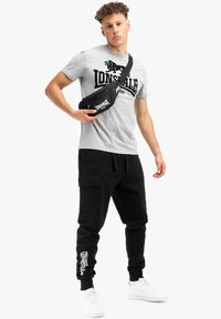 Grå t-shirt med svart "Lonsdale"-logga, svarta cargobyxor med ankelfickor och en svart axelväska. Vita sneakers fulländar looken.
