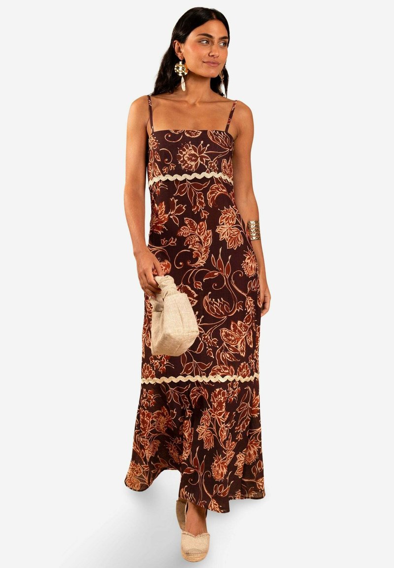 Mujer vestida con un maxi vestido floral marrón de tirantes finos, sosteniendo una bolsa de mimbre beige, y llevando pendientes grandes de oro y zapatos beige.