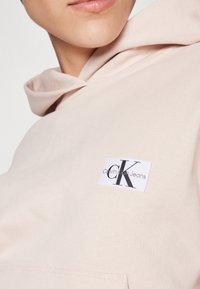 Gros plan d'une personne portant un sweat à capuche beige clair avec un patch logo Calvin Klein Jeans blanc sur la poitrine.
