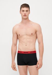 Fekete boxer alsónadrág piros gumis derékkal, rajta a "HUGO" logóval, sima anyagból készült, testhezálló kialakítással, férfi modellen bemutatva.