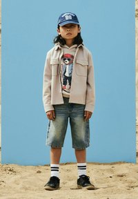 Jeune personne portant une casquette bleue, une veste beige, un sweat à capuche avec un motif d'ours, un short en jean, des chaussettes rayées et des baskets noires, debout sur du sable sous un ciel bleu.