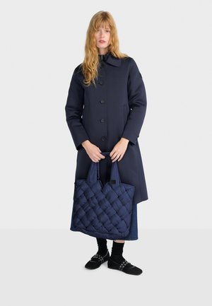 Cappotto lungo navy con colletto rotondo, bottoni frontali, abbinato a una borsa tote trapuntata navy e scarpe nere con cinghie decorative e calze.