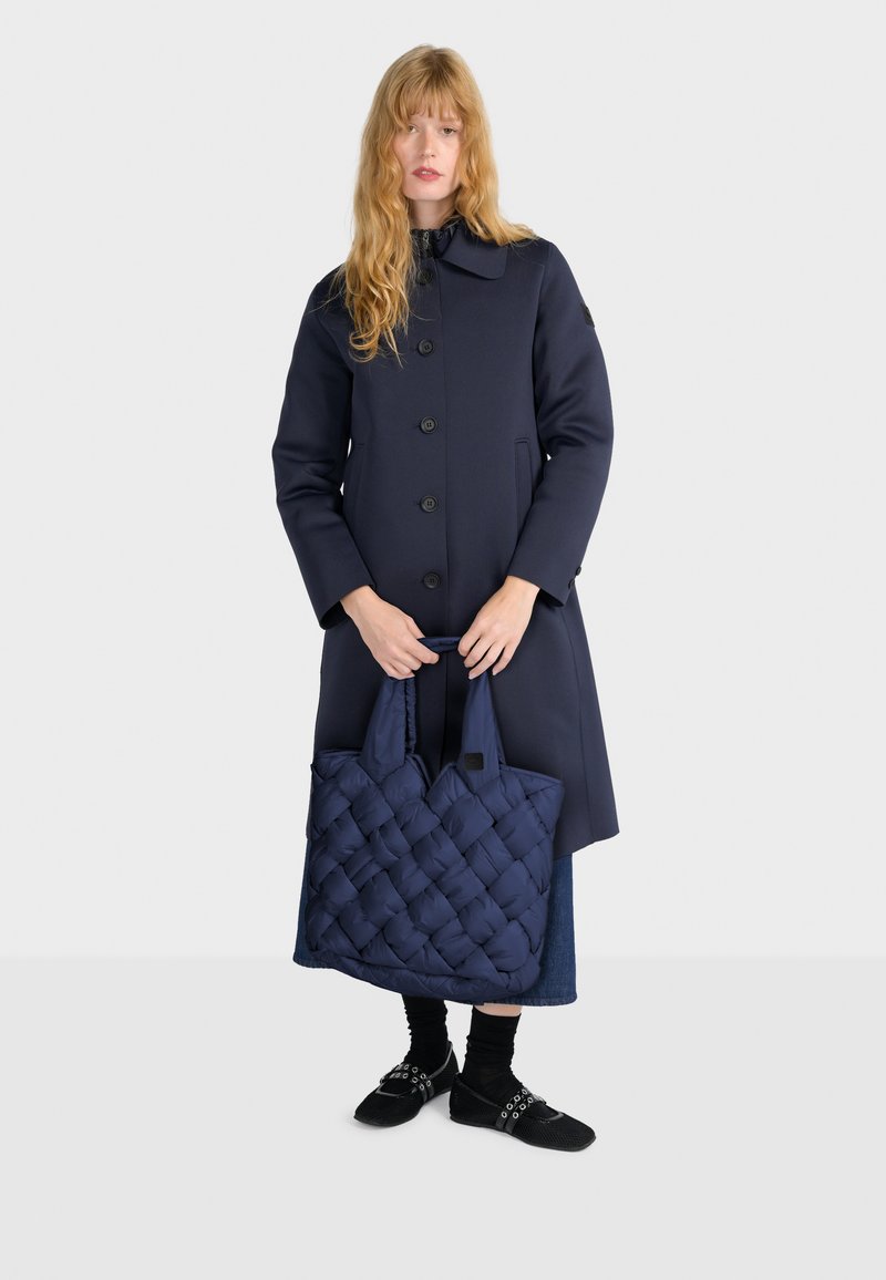 Marineblauwe lange jas met ronde kraag, voorknoopsluiting, gecombineerd met een gewatteerde marineblauwe tote bag en zwarte schoenen met decoratieve bandjes en sokken.