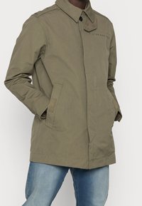 Giacca leggera verde oliva con colletto a bottoni, tasche frontali e dettagli di cucitura sottili. Presenta un design elegante e dritto.