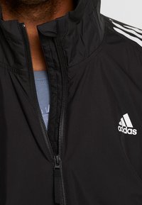 Svart zip-up jacka med hög krage, lätt material och vitt Adidas-logotyp på vänster bröst, med kontrasterande detaljer på ärmarna.
