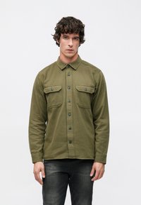 Olívazöld, gombos ing, pamut anyagból, hosszú ujjakkal, két mellkasi zsebbedugóval és szabványos gallérral. Sötét farmernadrággal viselve.