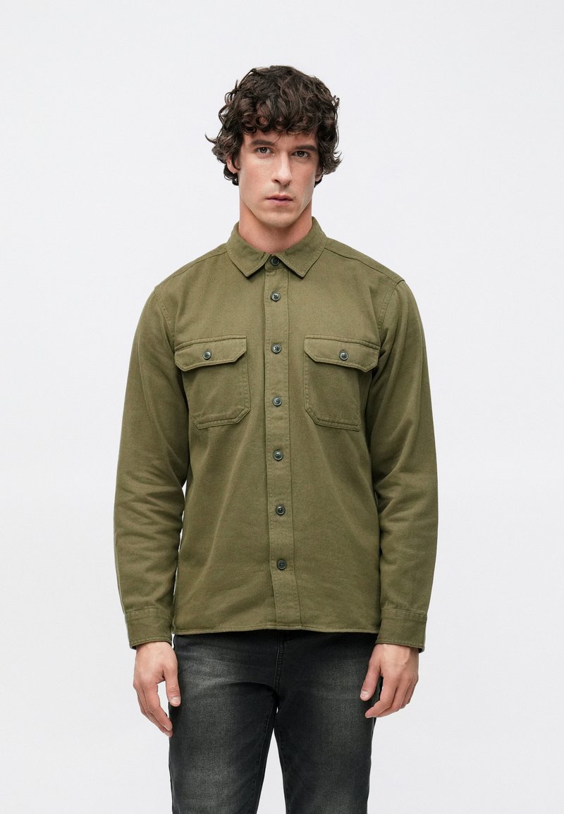 Olívazöld, gombos ing, pamut anyagból, hosszú ujjakkal, két mellkasi zsebbedugóval és szabványos gallérral. Sötét farmernadrággal viselve.