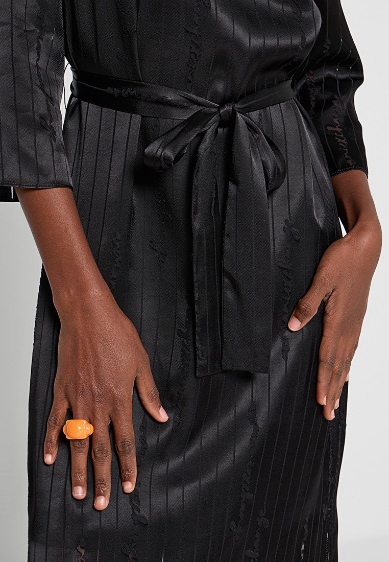 Černý saténový župan s vertikálními pruhy, uvázaný v pase odpovídajícím páskem. Má texturovaný vzor a na jedné ruce oranžový kroužek.