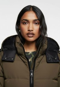 Pufferjacka med olivgrön kropp, svarta satinaccenter, dragkedja och tryckknappar, med hög krage och huva.