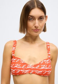 Pomarańczowy top bikini z szerokimi ramiączkami i ukształtowanym krojem, zdobiony białym, odręcznie pisanym wzorem logo na całej powierzchni. Gładka tekstura materiału.