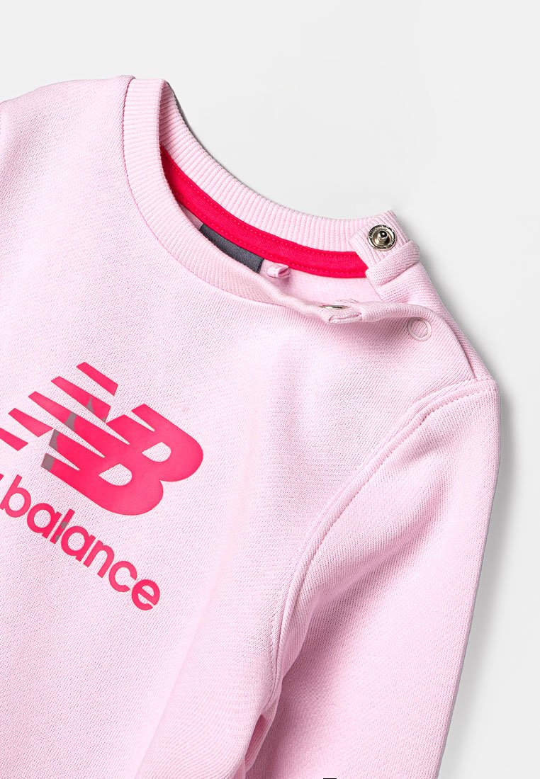 Sweatshirt rose clair avec col rond, présentant un logo "New Balance" rose vif, un détail de boutons à pression et une bordure de col rouge contrastante.
