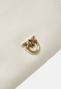 Balta tekstūruota odinė rankinė su auksinės spalvos metaliniu akcentu, kuriame yra du paukščiai ir apvalus žiedas. Lygus paviršius ir minimalistinis dizainas.