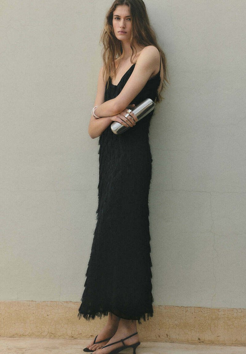 Femme en robe noire texturée sans manches tenant une pochette argentée, portant des bracelets et des sandales, debout contre un mur gris uni.