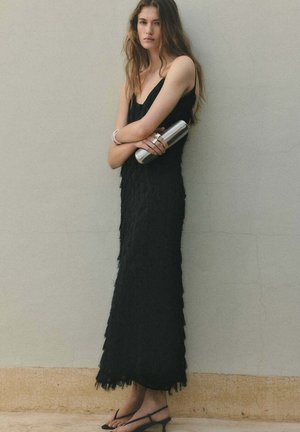 Mujer con un vestido negro sin mangas texturizado, sosteniendo un bolso de mano plateado, llevando pulseras y sandalias, de pie frente a una pared gris lisa.