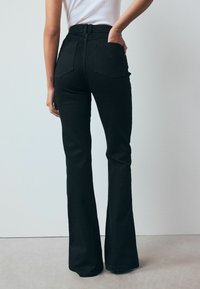Black flared jeans made of denim, high-waisted with a belt, featuring two rear pockets and smooth texture. No visible patterns or accents.