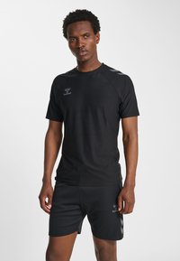 Man die staat en een zwart sportshirt met korte mouwen en bijpassende korte broek draagt met subtiele grijze logo's en chevron-details op een witte achtergrond.