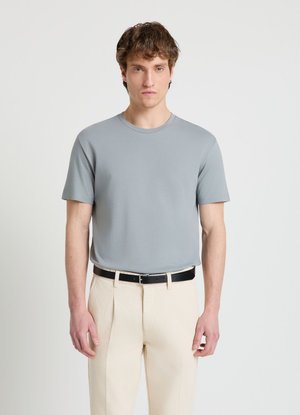 Giovane uomo con capelli corti e ricci castani indossa una camicia a maniche corte grigio chiaro, pantaloni beige e cintura nera, in piedi davanti a uno sfondo semplice.
