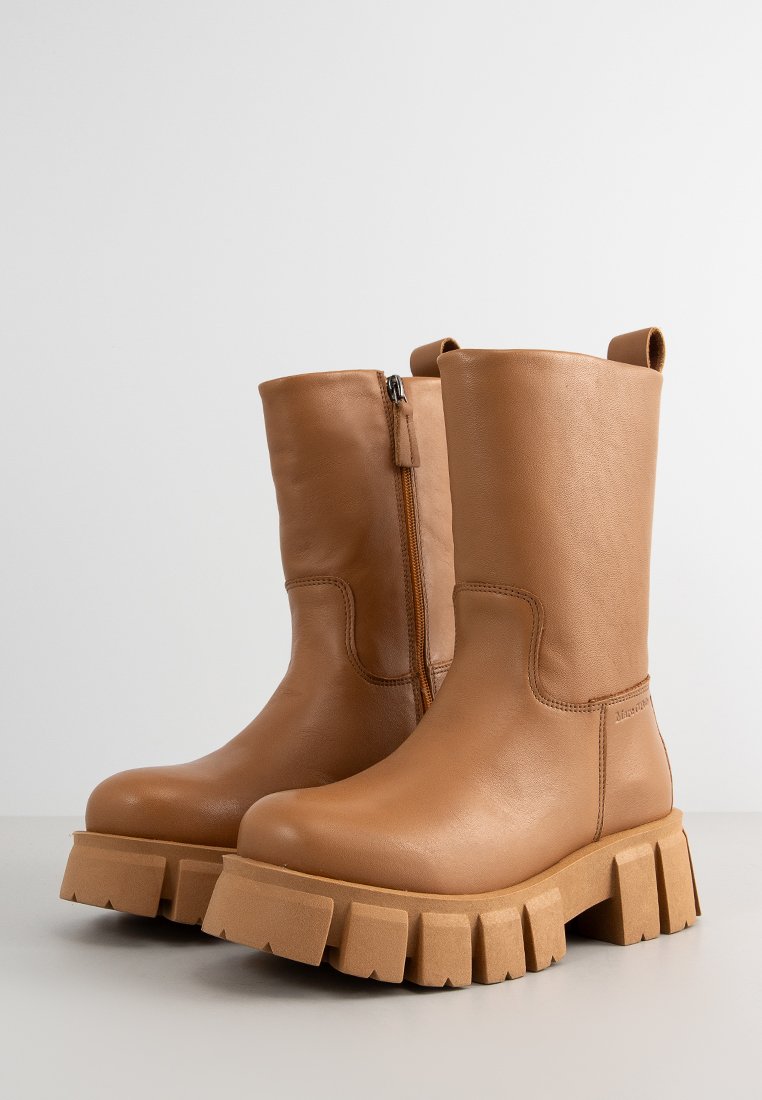 Ein Paar mittelbraune Lederstiefel bis zur Wade mit robusten Profilsohlen und seitlichen Reißverschlüssen auf einer hellgrauen Oberfläche vor einem weißen Hintergrund.