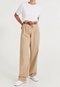 Pantalons beige taille haute à jambes larges avec plis sur le devant ; associés à un t-shirt blanc et une ceinture en cuir marron ; baskets blanches.