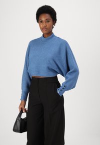 Maglione blu a coste, cropped, con collo alto e maniche ampie, dotato di bottoni sui polsini, abbinato a pantaloni neri a vita alta.