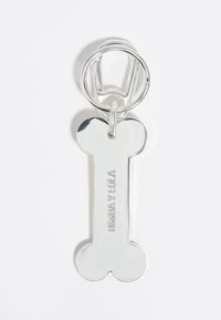 Portachiavi in metallo argentato a forma di osso, con testo in rilievo "BIMBA Y LOLA" e anello a gabbia per il fissaggio. Finitura superficiale liscia.