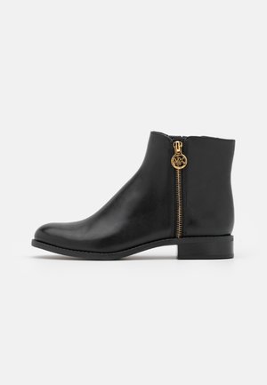 Bottine en cuir noir à talon bas avec fermeture éclair dorée et pendentif rond avec logo sur le côté, sur fond uni.