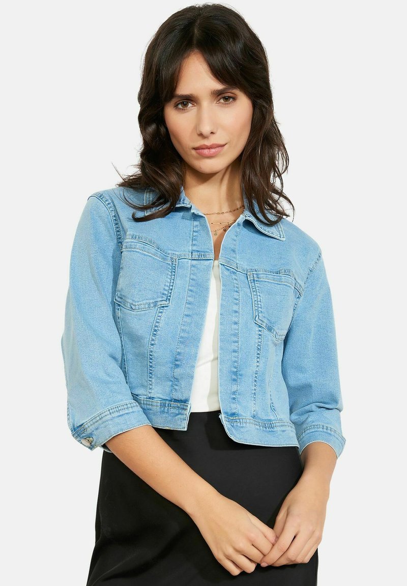 Mujer con una chaqueta vaquera corta azul claro sobre una camiseta blanca y una falda negra, de pie contra un fondo blanco liso.