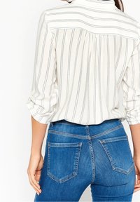 Vrouw in een wit shirt met zwarte verticale strepen en opgerolde mouwen, gecombineerd met blauwe denim jeans, gezien van achteren.