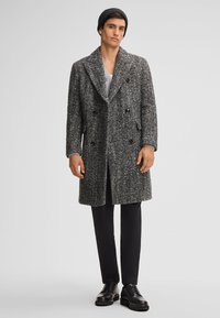 Man in een zwarte beanie, een grijze getextureerde dubbelzijdig geopende overjas, zwarte broek en zwarte leren schoenen, staande tegen een grijze achtergrond.