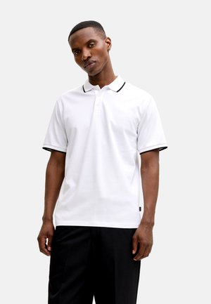 Weißes Poloshirt mit schwarzem Kragen und Ärmelbündchen, dreiknöpfiger Knopfleiste und dezentem Logodetail auf der Brust. Kombiniert mit schwarzer Hose.