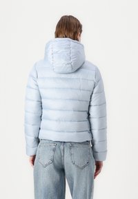 Polstret lyseblå jakke med hette, med horisontale quiltede linjer og en formsydd silhuett, kombinert med lyse vaskede jeans.