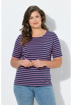 Vrouw met lang bruin haar die een marineblauw en roze gestreept shirt met korte mouwen en lichtblauwe spijkerbroek draagt, staand tegen een witte achtergrond.