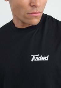 Melna kokvilnas T-kreklu ar baltu grafisko logo "Fadëd" kreisajā krūtīs, ar apaļu kakla izgriezumu un īsām piedurknēm.