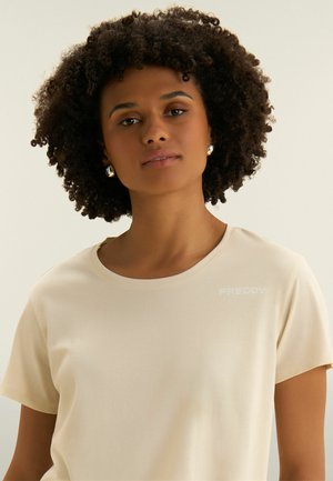 Mujer con cabello rizado que lleva una camiseta de manga corta de color beige claro con el logo "FREDDY" y pendientes de aro plateados, mirando hacia adelante sobre un fondo neutro.