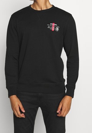 Homme portant un pull noir à manches longues avec un petit logo Diesel blanc et rouge ainsi qu'un texte sur le côté gauche de la poitrine, assorti d'un pantalon noir.