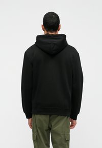 Μαύρα φούτερ με μεγαλη πίσω τσέπη και ρυθμιζόμενη κουκούλα, κατασκευασμένο από υφαντό ύφασμα. Συνδυάζεται με ελαιοπράσινα cargo παντελόνια.