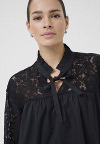 Sort bluse med lange ærmer i blonde, en volangkrave og en sløjfedetalje ved halsen. Glat stof med en løs pasform.